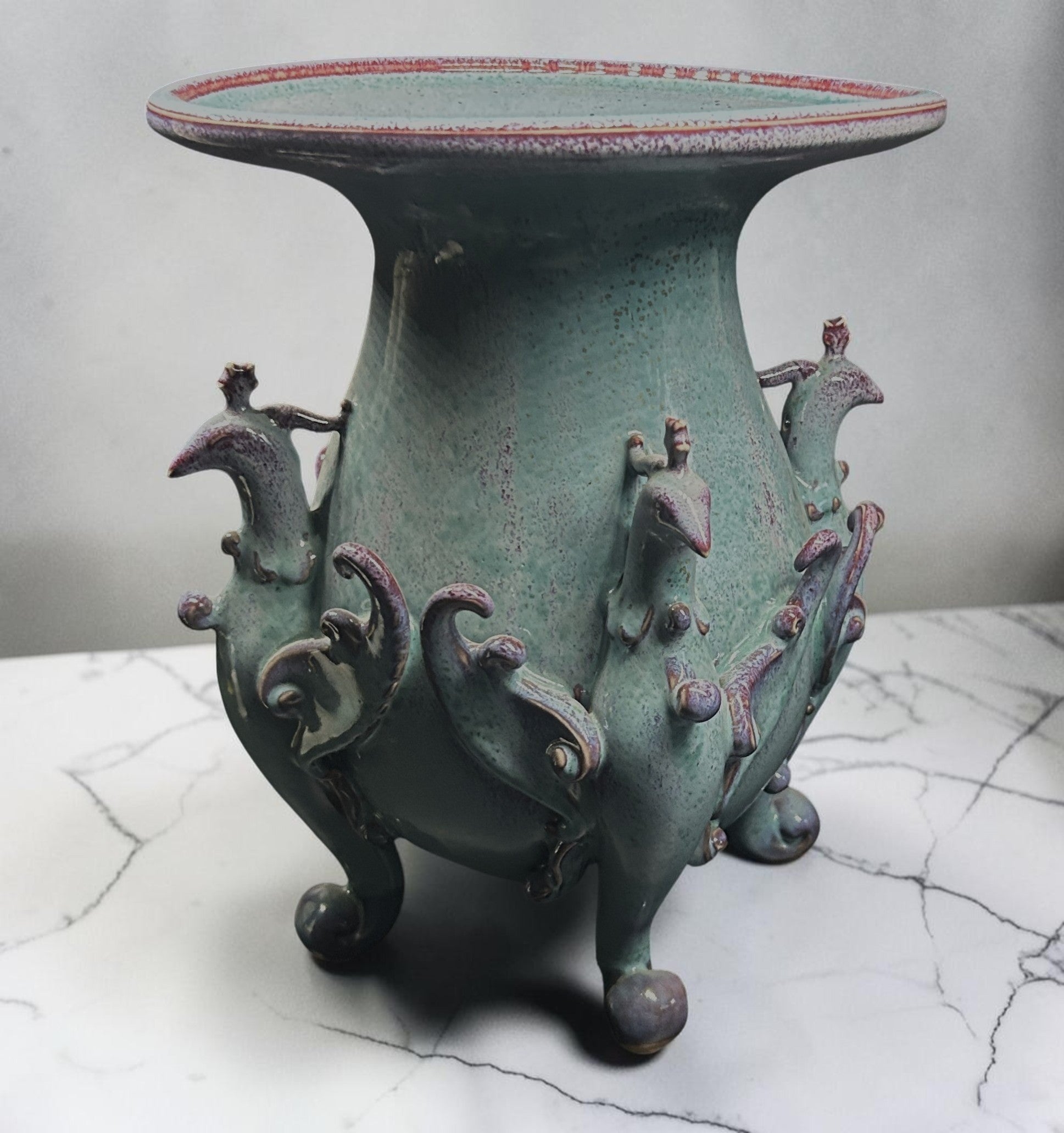 C&C Atelier celadon ceramic vase with purple accents, sculpted peacock motifs, and a tripod base. Ideal for floral arrangements or decor.